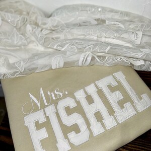 K&ouml;nnte beinhalten: Ein beiges Kissen mit dem Namen "Mrs. FISHEL" in wei&szlig;en, college-artigen Buchstaben gestickt. Zarter wei&szlig;er Spitzenstoff ist im Hintergrund drapiert, was auf ein Hochzeitsthema hindeutet.