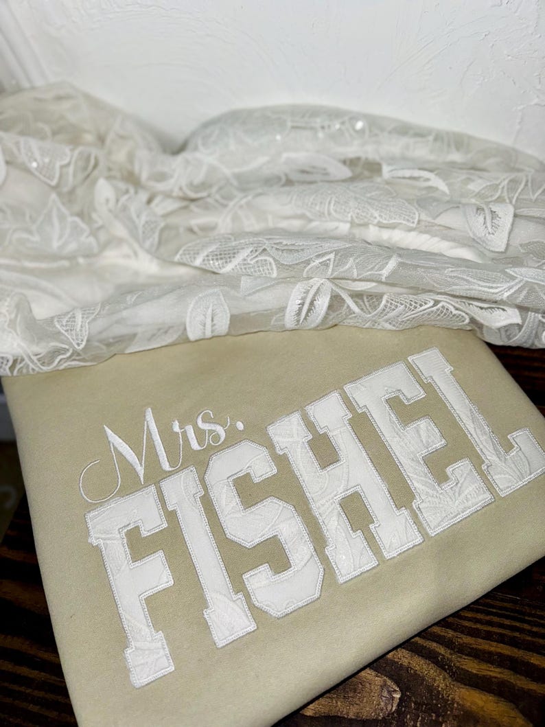 K&ouml;nnte beinhalten: Ein beiges Kissen mit dem Namen "Mrs. FISHEL" in wei&szlig;en, college-artigen Buchstaben gestickt. Zarter wei&szlig;er Spitzenstoff ist im Hintergrund drapiert, was auf ein Hochzeitsthema hindeutet.