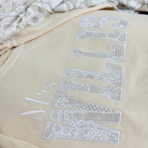 May include: A cream-coloured sweatshirt featuring the embroidered text "Mrs. ENTWINED" in white lace-like fabric. The letters are large and bold, with a delicate, textured appearance. A folded piece of lace fabric is in the background.