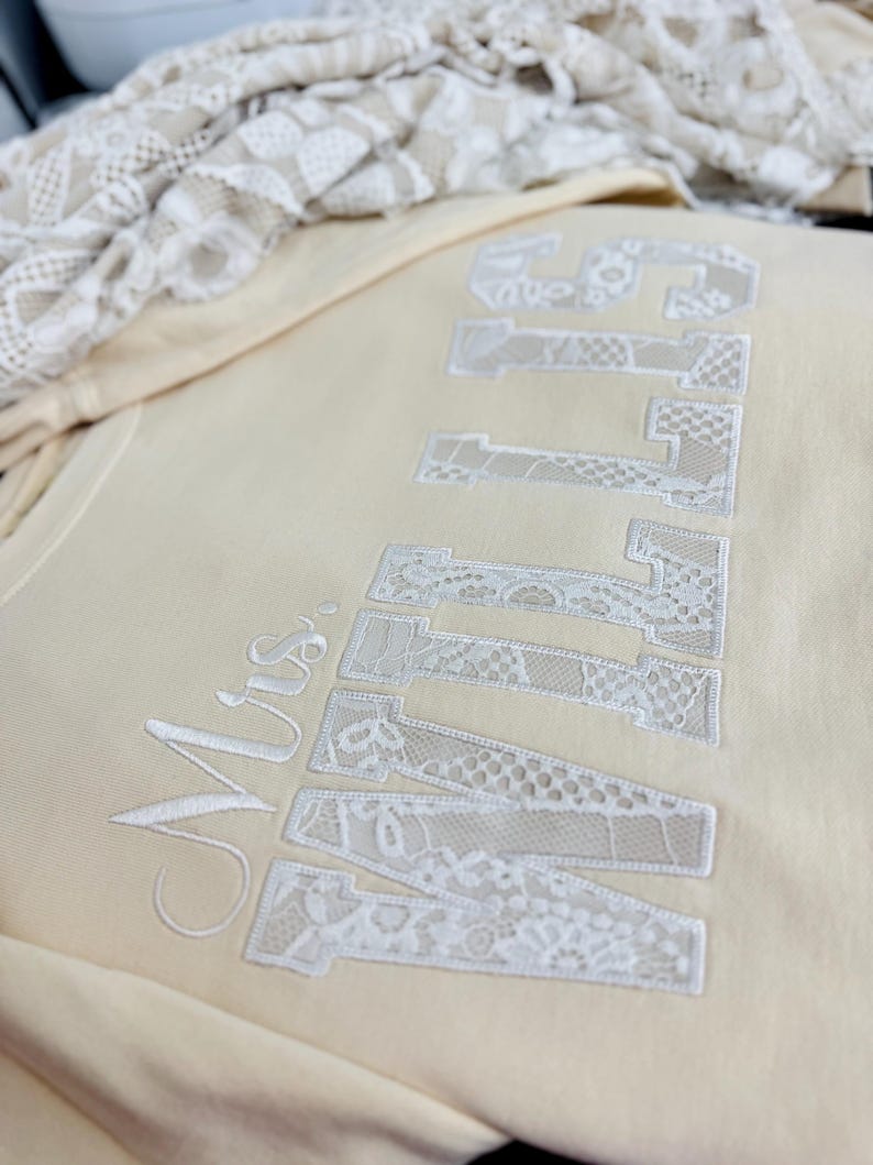 May include: A cream-coloured sweatshirt featuring the embroidered text "Mrs. ENTWINED" in white lace-like fabric. The letters are large and bold, with a delicate, textured appearance. A folded piece of lace fabric is in the background.