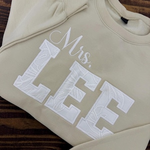 K&ouml;nnte beinhalten: Beiges Sweatshirt mit dem Namen "Mrs. LEE" in Wei&szlig; gestickt. Die Buchstaben sind gro&szlig; und blockartig, mit einem floralen Spitzenmuster, das die Buchstaben ausf&uuml;llt. Das Wort "Mrs." ist in Schreibschrift gehalten.
