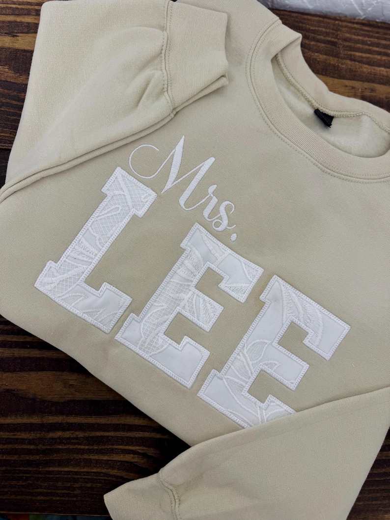K&ouml;nnte beinhalten: Beiges Sweatshirt mit dem Namen "Mrs. LEE" in Wei&szlig; gestickt. Die Buchstaben sind gro&szlig; und blockartig, mit einem floralen Spitzenmuster, das die Buchstaben ausf&uuml;llt. Das Wort "Mrs." ist in Schreibschrift gehalten.