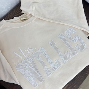 May include: A cream-coloured sweatshirt featuring the embroidered text "Mrs. WILLIS" in white. The letters are filled with a delicate lace pattern. The sweatshirt is folded on a dark wooden surface.