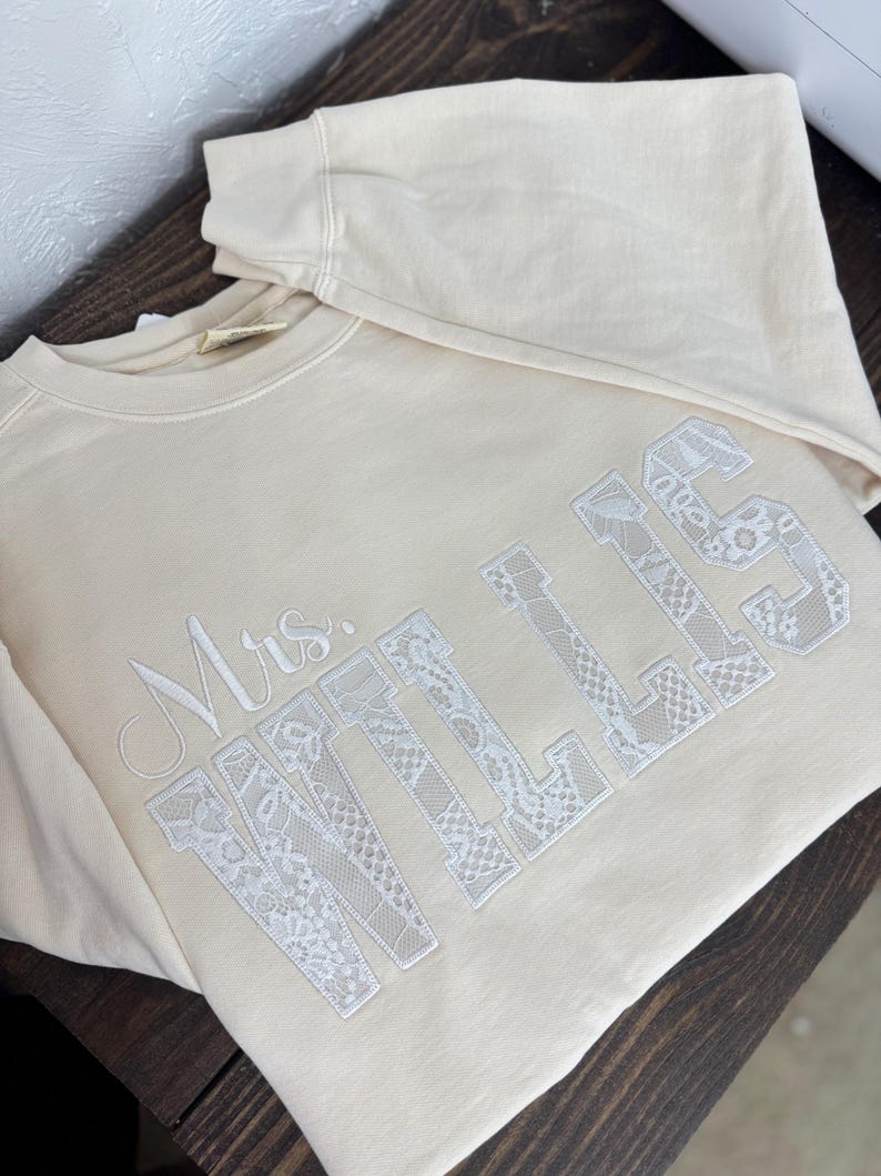 May include: A cream-coloured sweatshirt featuring the embroidered text "Mrs. WILLIS" in white. The letters are filled with a delicate lace pattern. The sweatshirt is folded on a dark wooden surface.