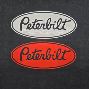 Op de afbeelding: Twee ovale Peterbilt logo stickers. De bovenste sticker heeft een witte achtergrond met zwarte tekst en een zwarte rand. De onderste sticker heeft een rode achtergrond met zwarte tekst en een zwarte rand. Beide stickers staan op een zwarte achtergrond.