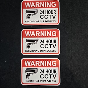 May include: Three rectangular warning stickers with rounded corners. Each sticker has a red top section with the word "WARNING" in white. Below, it reads "24 HOUR CCTV RECORDING IN PROGRESS" with a camera graphic.