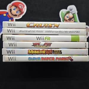 Puede incluir: Una pila de juegos de Wii, con títulos como "Cruis'n", "Fantastic Four", "Wii Fit", "Untamed", "Monster 4x4" y "Super Paper Mario". Las carátulas blancas muestran títulos coloridos. Pegatinas de Mario y Luigi en el fondo.