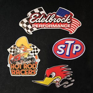 May include: Four automotive-themed stickers on a black background. The stickers include "Edelbrock Performance" with checkered and American flag accents, "STP" in a red oval, a cartoon hot rod racer, and a red bird with a yellow beak.