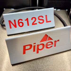 May include: Two rectangular, curved white signs with recessed red lettering. The top sign displays the text N612SL, while the bottom sign features the word Piper with a red triangular icon above the letter i. Both signs have a smooth, matte finish.