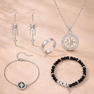 Könnte beinhalten: Eine Sammlung silberfarbener Schmuckstücke mit Stern- und Kompassmotiven. Enthält Ohrringe, einen Ring, eine Halskette und zwei Armbänder. Ein Armband hat schwarze Perlen und weiße Buchstabenperlen mit der Aufschrift "SKZ" mit einem Herzanhänger.