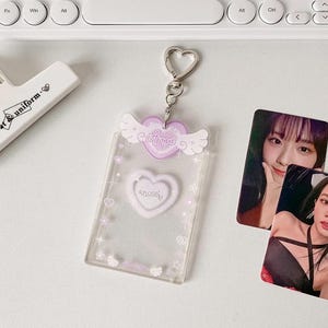 Pu&ograve; includere: Un portachiavi in plastica trasparente con un cuore e ali viola. Il cuore ha la scritta "Angel" su di esso. Il portachiavi ha una catena argentata e un anello a forma di cuore.