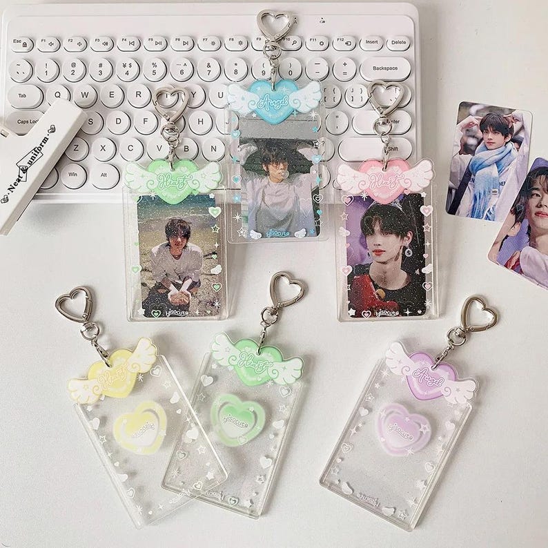 Pu&ograve; includere: Cinque portachiavi in plastica trasparente con un design a forma di cuore e ali. Ogni portachiavi ha un cuore e ali di un colore diverso. I portachiavi sono decorati con glitter e stelle. I portachiavi hanno un inserto fotografico in plastica trasparente. Gli inserti fotografici sono pieni di foto di persone. I portachiavi sono appesi a una superficie bianca.