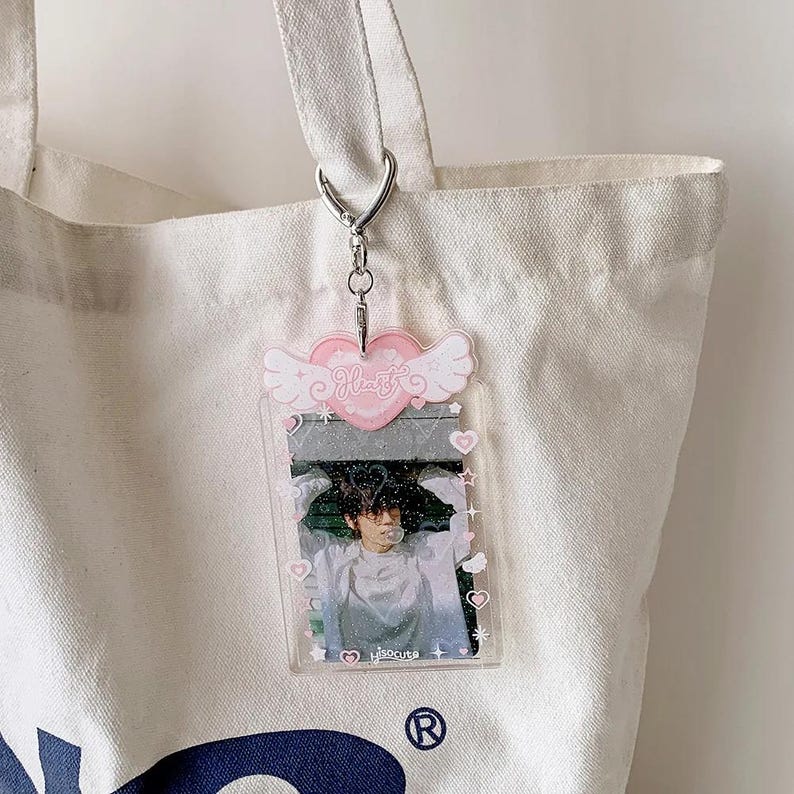 Pu&ograve; includere: Un portachiavi in plastica trasparente con un cuore rosa e ali che dice "Heart" e una foto di una persona all'interno. Il portachiavi &egrave; attaccato a una borsa tote bianca.