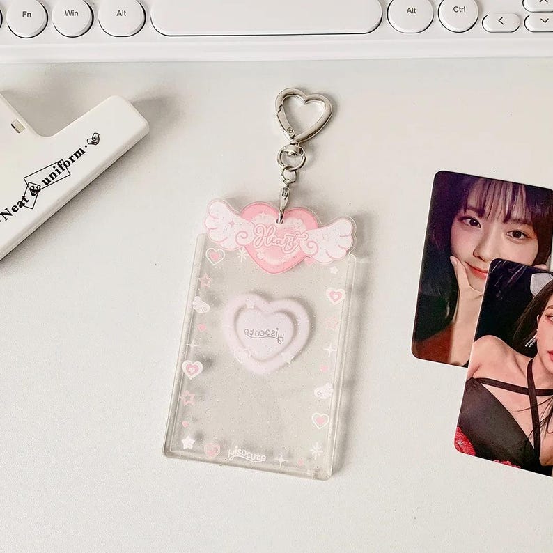 Pu&ograve; includere: Un portachiavi in plastica trasparente con un cuore rosa e ali. Il portachiavi ha un anello in metallo argentato e un inserto in plastica trasparente per una foto. Il testo "Hello" &egrave; stampato sul cuore. Il portachiavi &egrave; su una superficie bianca.