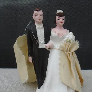 May include: A vintage wedding cake topper depicting a bride and groom. The groom is in a black tuxedo with a white bow tie, and the bride wears a white dress and holds a bouquet. The figures are painted with fine details and stand together.