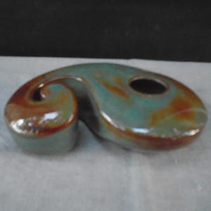 May include: A ceramic candle holder with a curved, abstract shape featuring a glossy glaze in blended shades of teal, green, and brown. The object has a smooth, rounded surface with a circular opening on one side for holding a candle.