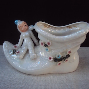 Könnte beinhalten: Ein Vintage-Pflanzgefäß in Schuhform aus Keramik mit einem skurrilen Design. Es hat eine weiße Oberfläche mit schillerndem Glanz, verziert mit kleinen floralen Akzenten in Rosa, Blau und Schwarz. Eine Figur eines Kindes in einem weißen Overall und einer blauen Mütze lehnt an dem Schuh.