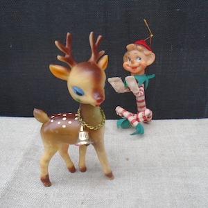 May include: Vintage Christmas decorations featuring a brown reindeer with white spots and a gold bell necklace, and a small elf with a red and white striped outfit. The reindeer has blue eyes and brown antlers. The elf is holding a scroll.