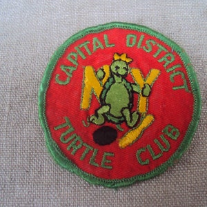 May include: A red, green, and yellow embroidered patch with a cartoon turtle wearing a crown and holding a bowling pin. The text on the patch reads "Capital District Turtle Club".