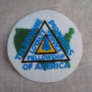 Puede incluir: Un parche circular con el texto "TELEPHONE PIONEERS OF AMERICA" en azul, rodeando un triángulo negro con las palabras "LOYALTY SERVICE FELLOWSHIP". Un mapa verde de los Estados Unidos está en el lado izquierdo del parche.
