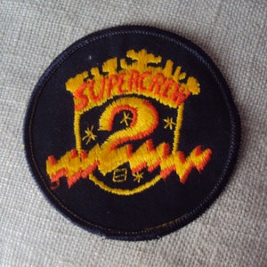 May include: A black embroidered patch with a yellow and orange design. The patch features the text "Supercrew" and a large number "2" with stars around it.