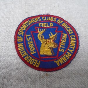 May include: A circular embroidered patch featuring a blue background with a red border. The center displays a gold deer head graphic. Text reads: FEDERATION OF SPORTSMENS CLUBS OF BUCKS COUNTY PENNA, FIELD, FOREST, and STREAM in gold lettering.