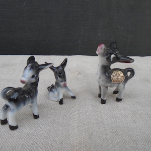 May include: Three ceramic donkey figurines. The donkeys are black, white, and gray with pink accents on their ears and noses. One donkey is standing, one is sitting, and one has a gold emblem on its side. The figurines are on a light-colored surface.