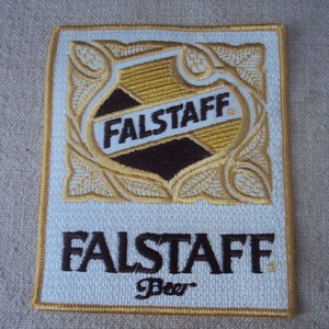 May include: A rectangular embroidered patch featuring a white background with a gold border. The design includes a central shield with the text FALSTAFF in dark brown, surrounded by gold leaf patterns. Below the shield, the text FALSTAFF Beer is embroidered in dark brown.