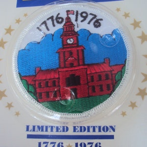 Puede incluir: Un parche redondo blanco bordado con un edificio rojo con una torre del reloj y una pequeña bandera estadounidense en la parte superior. El edificio está rodeado de nubes azules y hierba verde. El texto "1776 1976" está escrito en blanco en la parte superior del parche. El texto "LIMITED EDITION 1776 1976" está escrito en negro sobre un fondo blanco debajo del parche.