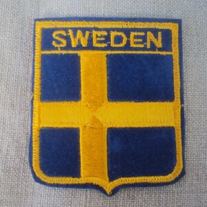 Könnte beinhalten: Ein blauer und gelber gestickter Aufnäher mit dem Wort "SWEDEN" in gelben Buchstaben. Der Aufnäher zeigt die schwedische Flagge, ein gelbes Kreuz auf blauem Hintergrund.