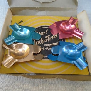 Può includere: Un set di quattro posacenere individuali in metallo nei colori blu, rosa, oro e verde acqua, ciascuno con una coppa centrale e tre tacche. Sono disposti all'interno di una scatola di cartone con un inserto giallo con la scritta Tach-a-Tray Individual Ash Trays.