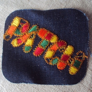 May include: A blue denim patch with a colorful embroidered design. The design features a series of interlocking shapes in shades of red, orange, yellow, green, and gold.