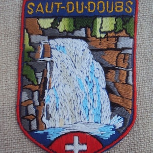 Puede incluir: Parche bordado con fondo azul y borde rojo. El parche presenta una cascada con un diseño blanco y azul, un bosque verde y una formación rocosa marrón. El texto "SAUT-DU-DOUBS" está bordado en amarillo en la parte superior del parche. Un círculo rojo con una cruz blanca se encuentra en la parte inferior central del parche.