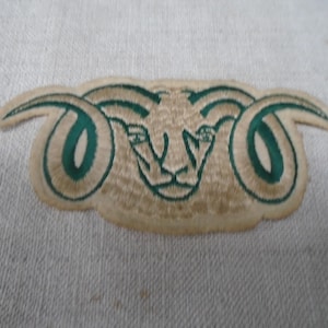Op de afbeelding: Een geborduurde patch met een ontwerp van een ramshoofd. De ram is uitgevoerd in lichtbruin garen met donkergroene accenten die de grote gekrulde hoorns en de gelaatstrekken omlijnen. De patch heeft een vorm die de contouren van de ram volgt.