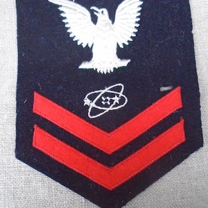 May include: A navy blue patch with a white eagle emblem above two red chevrons. A white design with stars and a circular shape is below the eagle. This patch is likely a military rank insignia.