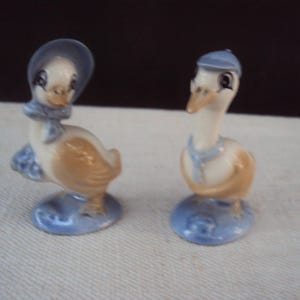 May include: Two ceramic figurines of geese wearing blue hats and scarves. The goose on the left is wearing a bonnet and the goose on the right is wearing a cap. Both geese are standing on a blue base.