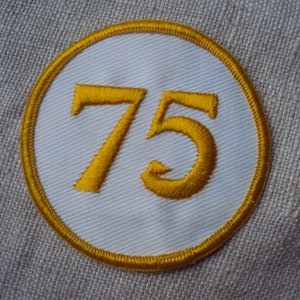 May include: A white circular patch with a yellow embroidered border and the number 75 in yellow.