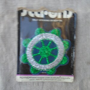 May include: A green and silver embroidered patch in a wheel design, on a black backing. The patch is in a clear plastic sleeve. The text "SELF-STICKING OBSESSION" is visible at the top.