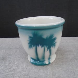 May include: White ceramic egg cup with a teal palm tree design. The egg cup has a flared shape with a teal rim and base. The palm trees are silhouetted against the white background.