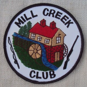 May include: White embroidered patch with black trim, featuring a red-roofed house with a water wheel, green trees, and a blue stream. The text "MILL CREEK CLUB" is written in black around the image.