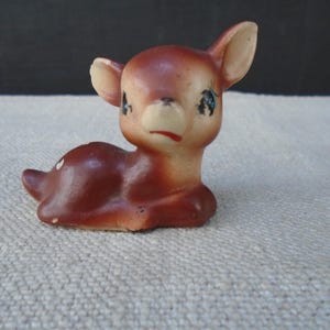 May include: A small ceramic figurine of a resting fawn with a brown body, a cream-colored face, and large ears. The deer has painted blue eyes with long eyelashes, a small black nose, and a red mouth, with white spots visible on its back.