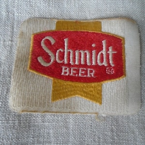 May include: A vintage Schmidt Beer patch. The patch features a red and gold logo with the word "Schmidt" in white script above "BEER". The patch is rectangular with a white background and gold accents.