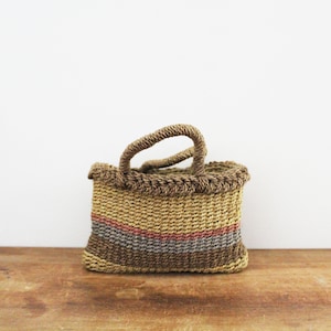 Może przedstawiać: Mały, tkany kosz z uchwytem. Kosz jest prostokątny z beżowym, brązowym i czerwonym wzorem w paski. Uchwyt i obręcz są ciemnobrązowe. Kosz stoi na drewnianej powierzchni.