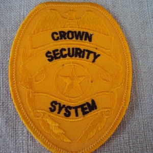 May include: Yellow embroidered patch with a black outline. The patch features a stylised eagle with wings spread, a star, and the text "CROWN SECURITY SYSTEM".