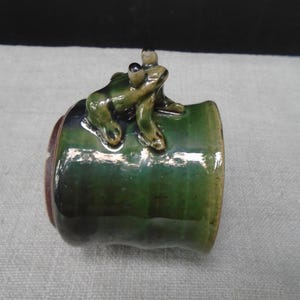 May include: A cylindrical ceramic mug featuring a glossy dark green glaze with a sculpted three-dimensional frog figure attached to the exterior. The frog is painted in shades of olive and brown, with prominent eyes and textured skin details.