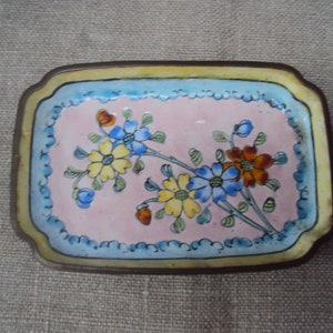 Può includere: Un vassoio rettangolare per bigiotteria con interno rosa, bordo azzurro e bordo giallo. Il vassoio presenta un motivo floreale con fiori blu, gialli e arancioni. Il vassoio ha una cornice in metallo color ottone.