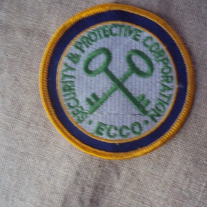 Peut inclure: Un écusson rond brodé de couleur jaune, bleu et vert avec le texte "Security & Protective Corporation, ECCO." et une paire de clés croisées au centre.