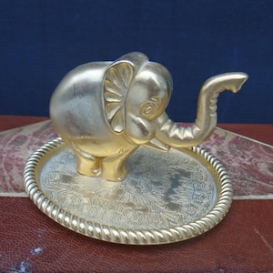 May include: A gold-coloured elephant figurine with an upturned trunk, standing on an oval tray. The tray has a floral design and a twisted rope-like border. The figurine and tray are made of metal.