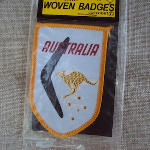 May include: A white woven patch with an orange border. The patch features a kangaroo and a boomerang in gold thread. The text "AUSTRALIA" is in red thread. The patch is packaged in a clear plastic bag with a yellow and brown header card that reads "SOUVENIR WOVEN BADGES COPYRIGHT ©".