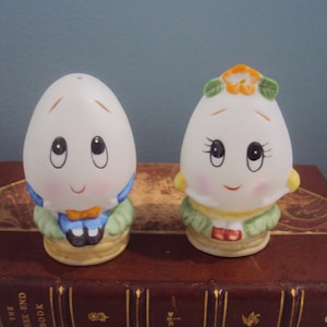 May include: A pair of ceramic salt and pepper shakers shaped like eggs with painted faces. The salt shaker is a blue egg with a bow tie and the pepper shaker is a yellow egg with a flower on its head.
